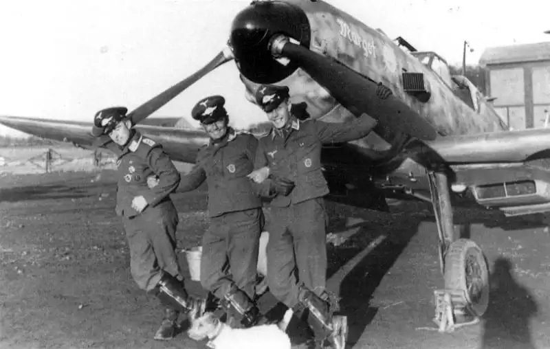 Three German fighter pilots posing beside an early Messerschmitt Bf 109 fighter aircraft named "Margot"