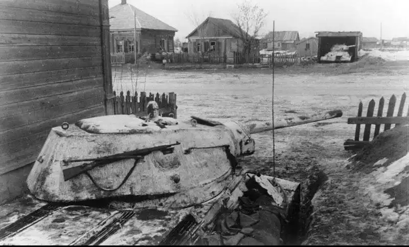 A winter-camouflaged Soviet T-34 tank with a captured German Kar98k rifle resting on its turret, dug into a trench in a village near Rzhev, 1942.