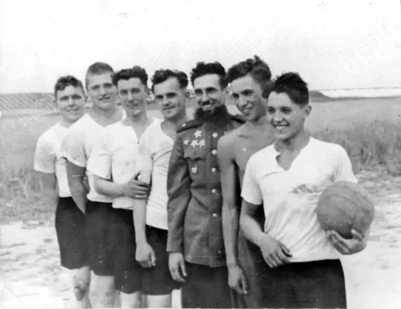 Black and white historical photo showing seven smiling Soviet Red Army soldiers and officers in sportswear holding a volleyball in July 1945.