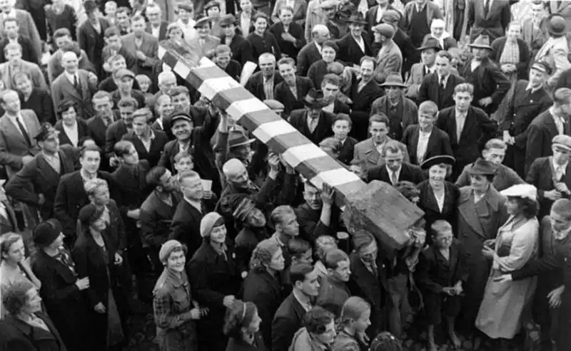 Black and white historical photo showing a dense crowd of people carrying a striped border pole on their shoulders in triumph during October 1938.