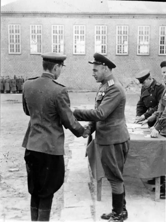 Black and white historical photo showing a Soviet Red Army officer shaking hands and awarding a medal to another officer in front of a brick building in July 1945.