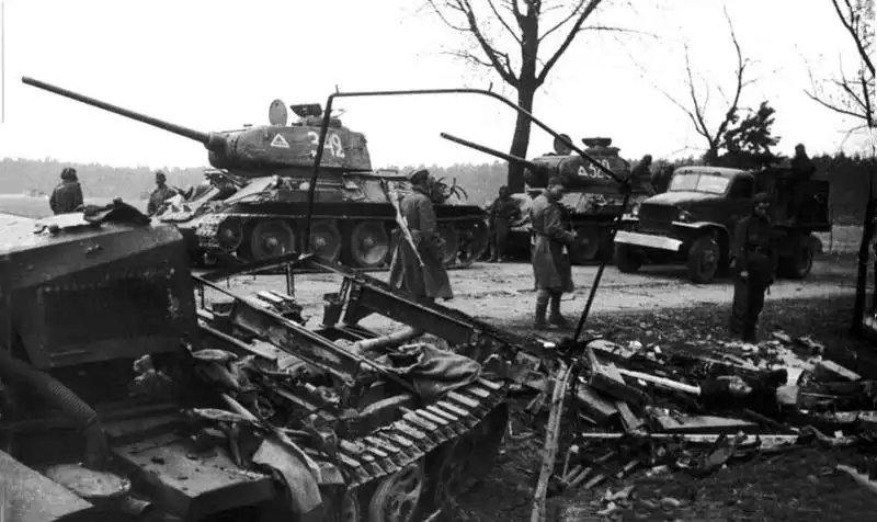 Two T-34-85 tanks of the 9th Tank Corps drive past a destroyed German Sd.Kfz. 11 halftrack and a Lend-Lease Chevrolet G7107 truck during the Battle of Berlin