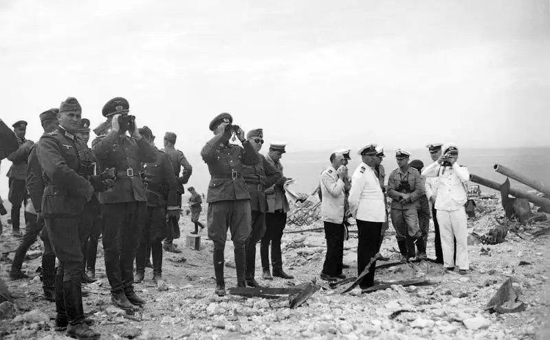Black and white historical photo showing German and Italian military officers standing among the concrete rubble and broken gun barrels of the 35th Coastal Battery in Sevastopol, 1942.