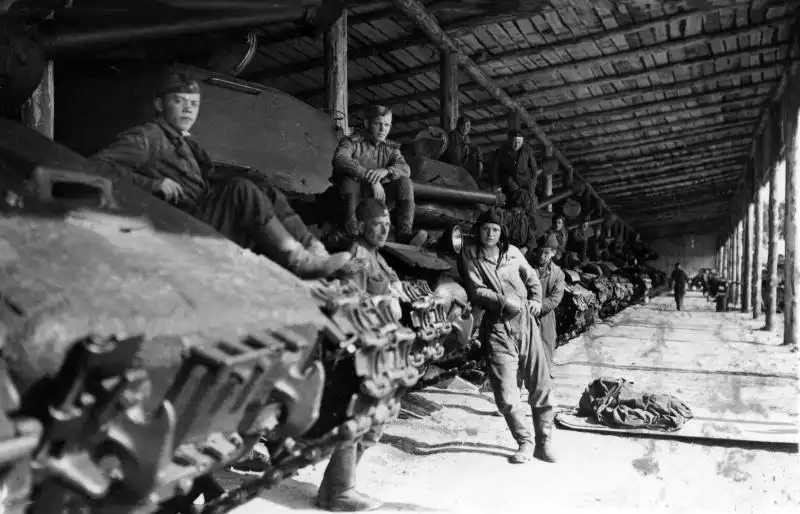 Black and white historical photo showing Soviet tank crews sitting and standing on a line of IS-2 heavy tanks inside a wooden shed in Güstrow, 1945.