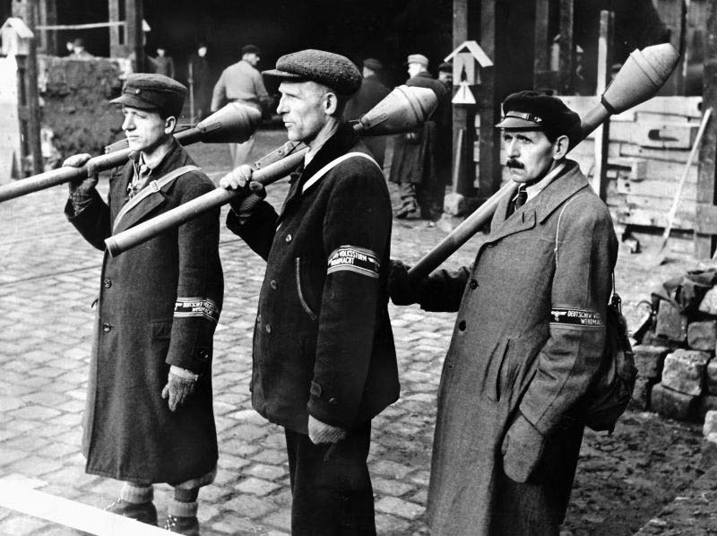 Black and white archival photo showing three elderly German civilian-clothed Volkssturm men wearing armbands and carrying Faustpatrone anti-tank weapons on a Berlin street, March 1945.