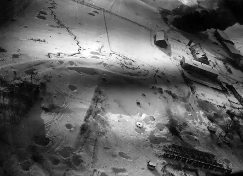 Top-down low-altitude aerial photo of a snow-covered village, zig-zag trenches, bomb craters, and a burning fuel depot in Grünwiese, East Prussia, in March 1945.