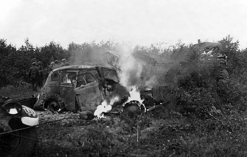 Black and white historical photo showing a burning German Opel Super 6 car in the foreground and a flipped-over Soviet T-34 tank with tracks visible in the background near Vilnius, June 1941.