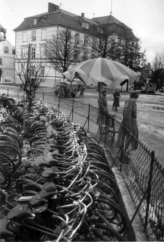Black and white historical photo showing long rows of captured bicycles lined up in a fenced town square guarded by Soviet soldiers in Bischofsburg, 1944.