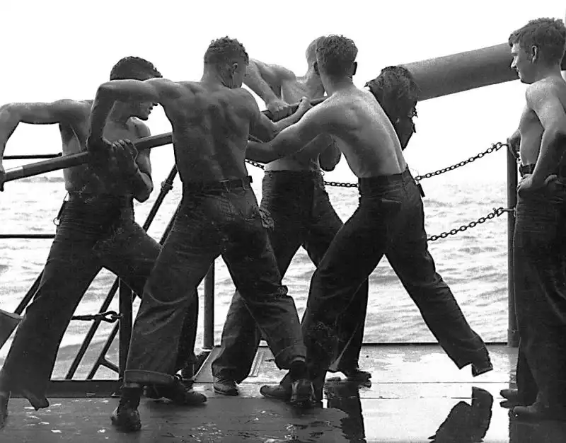 Black and white historical photo showing bare-chested US Navy sailors working together to clean a large naval gun barrel on a warship deck in the Pacific, 1945.