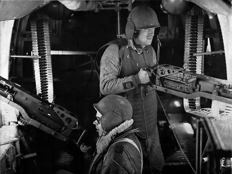 Black and white photo of two US bomber gunners wearing helmets and flak jackets inside a B-24 Liberator aircraft operating heavy machine guns in 1943.