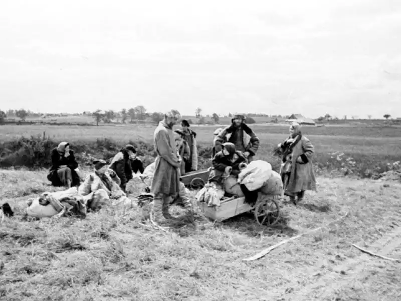 Black and white historical photo of Soviet rural refugees sitting on a field embankment with handcarts packed with belongings near Pskov in 1941.