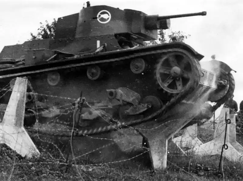Black and white WWII-era photo of a Polish 7TP light tank with a bison emblem on the turret crushing barbed wire and concrete dragon's teeth obstacles in 1938.