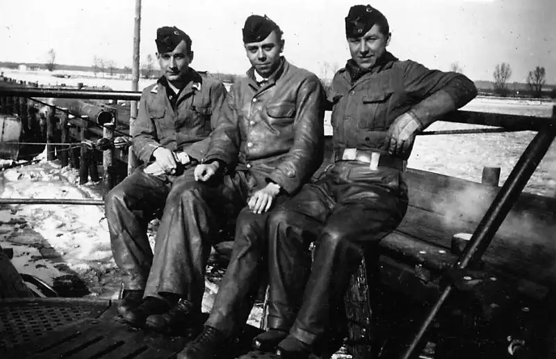 Black and white historical photo showing three German U-boat crew members sitting on the wooden deck of a submarine in spring 1944.
