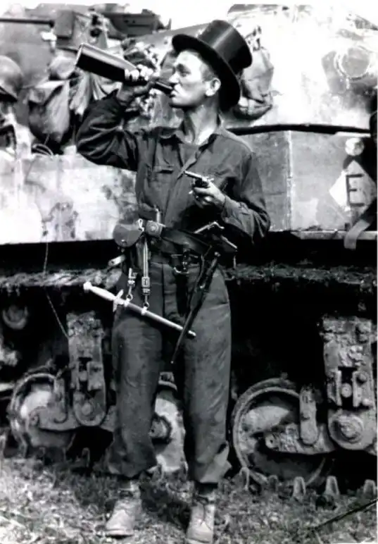 U.S. soldier wearing a top hat drinking from a bottle of French wine, adorned with captured German daggers, knives, pistols, and belt in Normandy 1944.