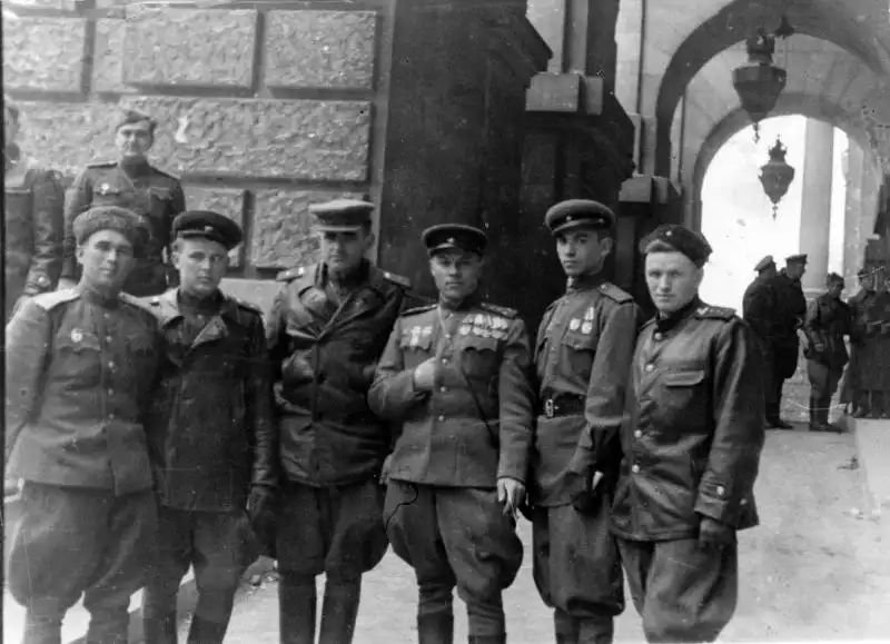 Black and white historical photo showing Soviet tank officers in uniforms and leather coats posing near the rusticated stone walls and archway of the Reichstag in Berlin, May 9, 1945.