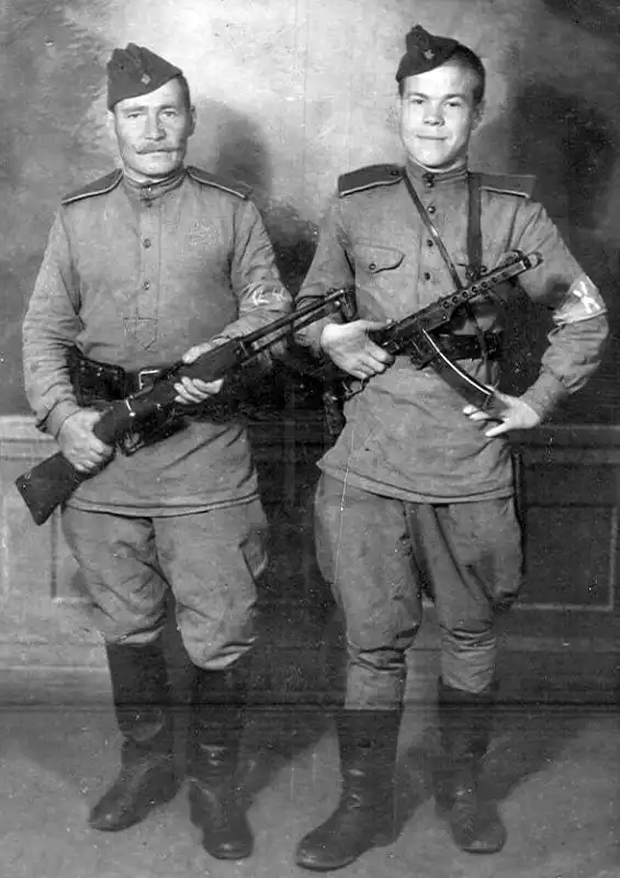 Two Soviet soldiers in military uniforms with armband insignias posing with a PPS-43 submachine gun and an Italian Mannlicher-Carcano folding bayonet carbine.