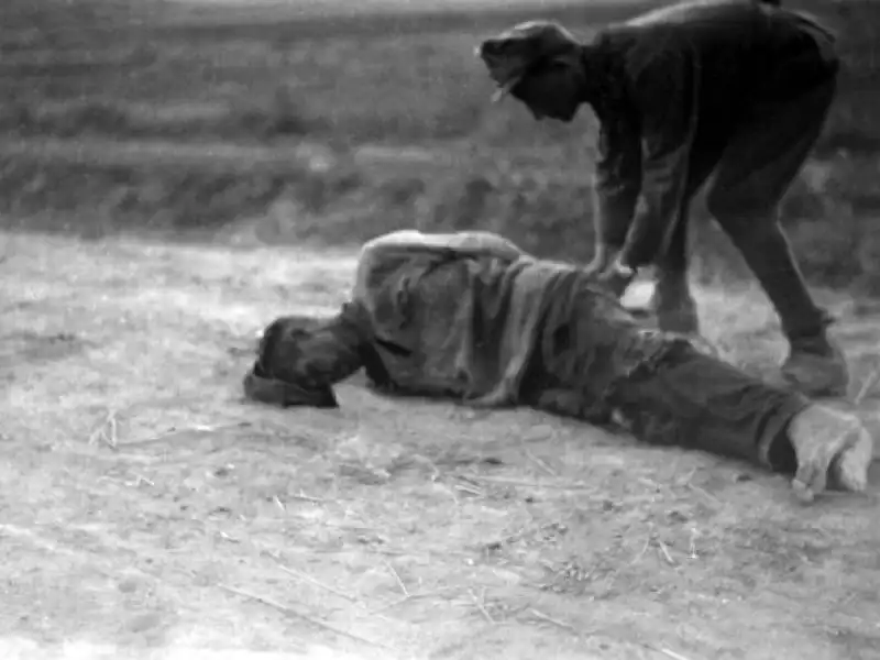 Black and white historical photograph of a Romanian soldier in field uniform leaning over a captured Soviet soldier lying on a dirt road in Crimea in 1942.