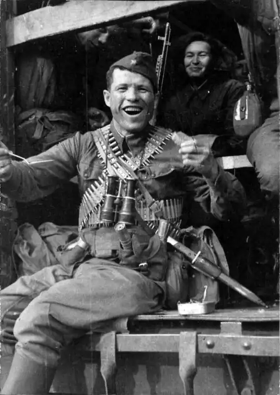 Smiling Soviet Red Army soldier sitting in a train car door, wearing ammo belts, holding French binoculars, with SVT-40 rifle and Mauser bayonet.