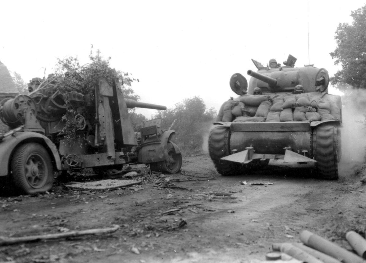 An American M4A3 Sherman tank drives past an abandoned German 88mm FlaK 36/37